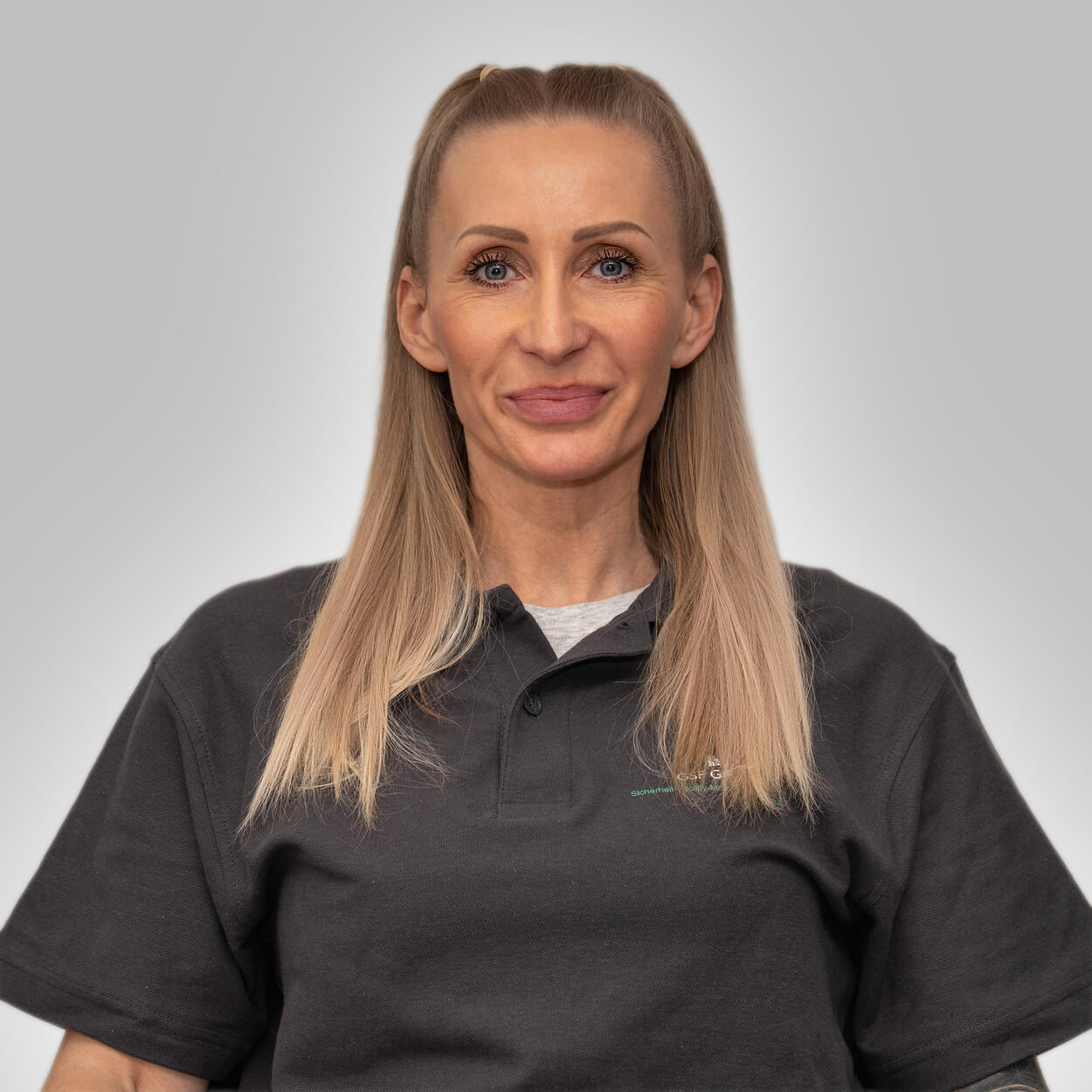 Eine blonde Frau mit neutralem Gesichtsausdruck trägt ein dunkelgraues Poloshirt