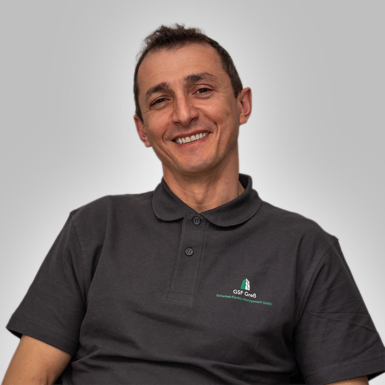 Ein lächelnder Mann mit kurzen braunen Haaren trägt ein dunkelgraues Poloshirt mit Logo