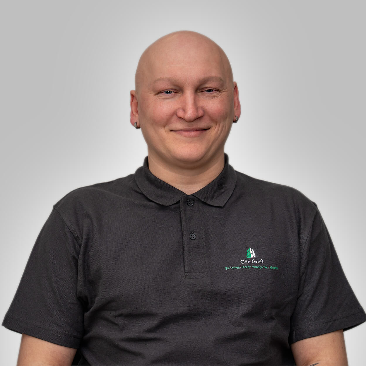 Ein glatzköpfiger Mann mit Ohrringen lächelt freundlich in schwarzem Poloshirt