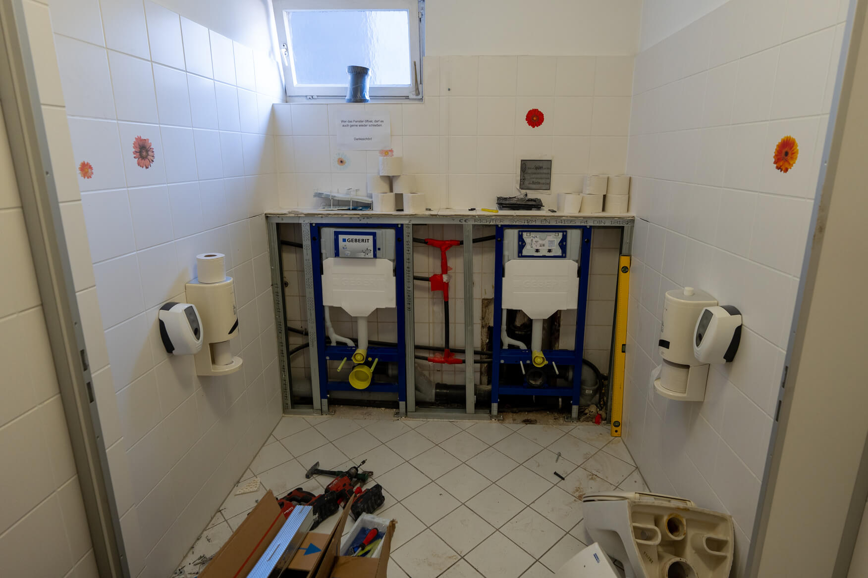 Zwei Toilettenrahmen ohne Verkleidung in einem weißen gefliesten Raum mit Werkzeug