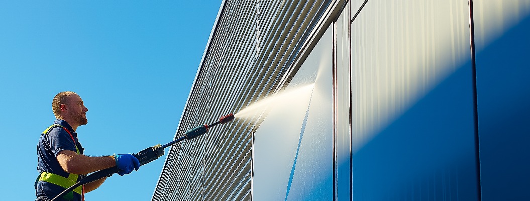 A worker is using a pressure washer to clean the facade of a modern commercial building