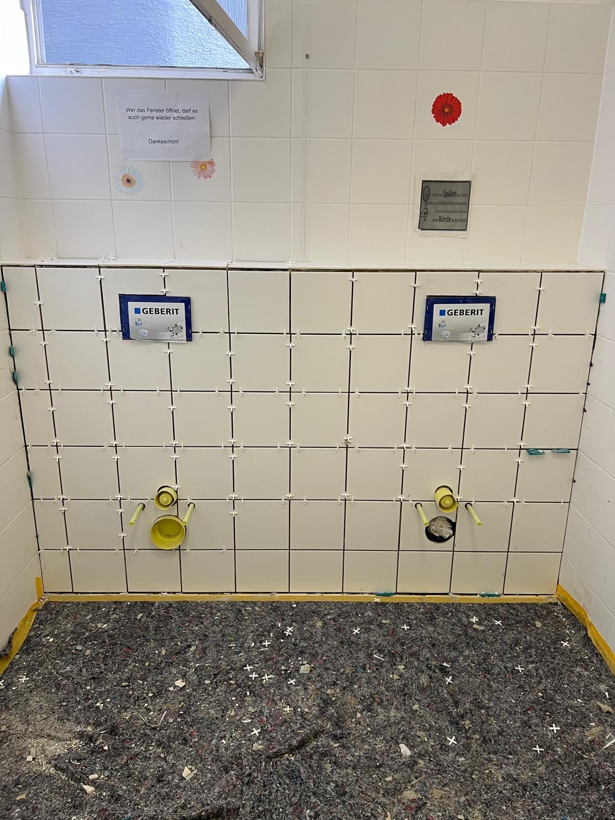 Eine halbfertige weiße Badezimmerwand mit sichtbaren Rohren und unfertigem Bodenbelag