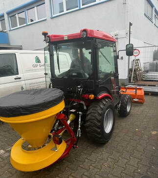 Eine Person mit neutralem Gesichtsausdruck sitzt in einem kleinen roten Traktor mit gelbem Streugerät