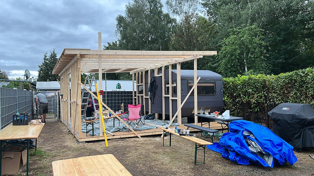 Holzunterstand im Bau neben grauem Wohnwagen mit blauer Plane und Biertischen