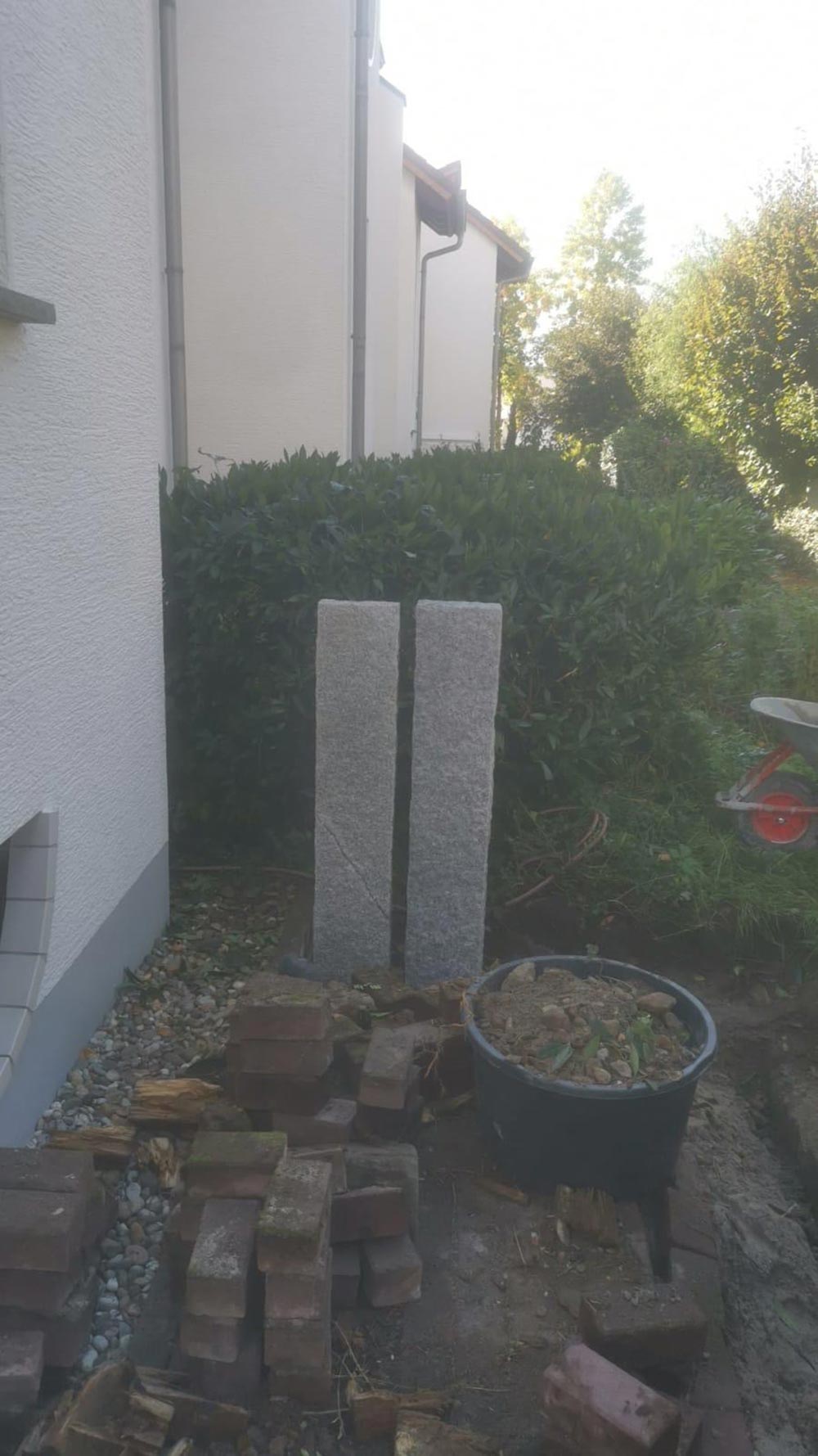Hof mit zwei grauen Steinpfosten neben Busch und roter Schubkarre