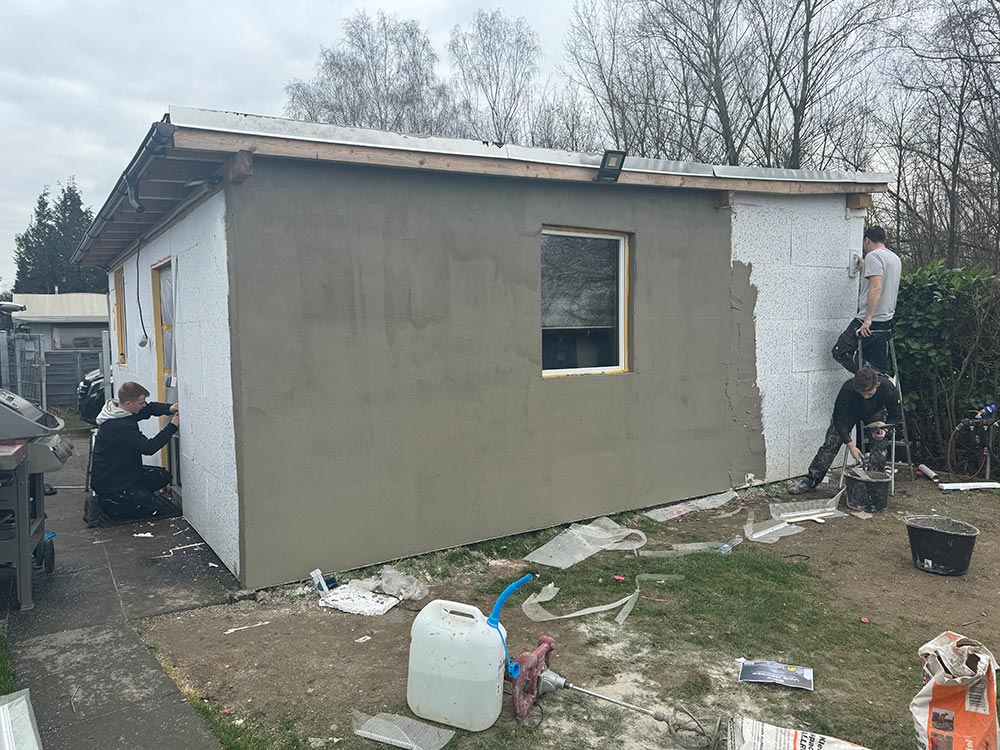 Zwei Maenner konzentriert beim Verputzen eines grauen Gartenhauses mit Fenster