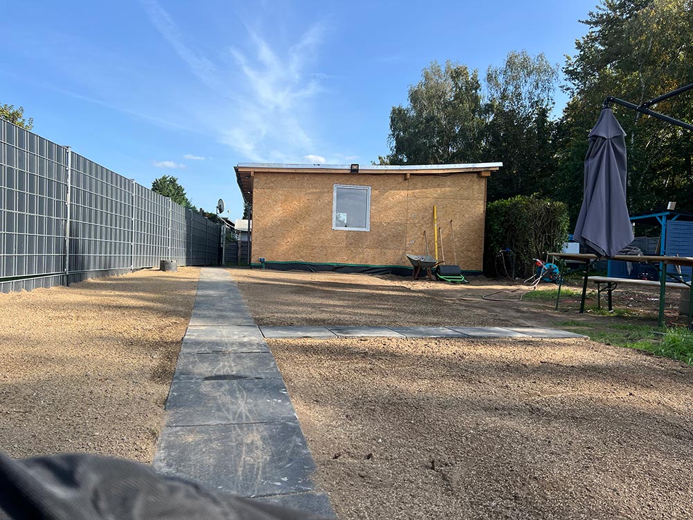 Garten mit braunem Haus und Fenster, Kiesweg, Schubkarre und Sonnenschirm