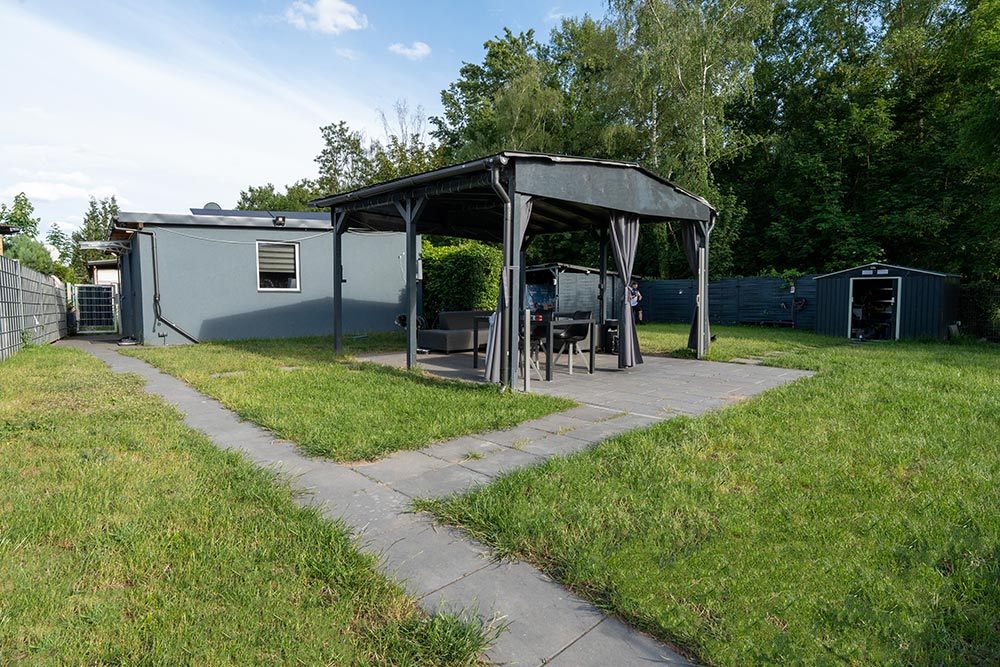 Graues Gartenhaus mit schwarzer Pergola und Sitzgruppe auf grüner Wiese