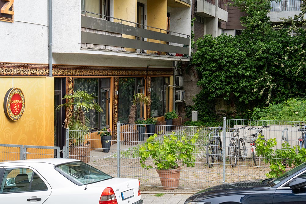 Gelbes Restaurant mit Glasfront und Pflanzen, daneben Fahrradzaun und Autos