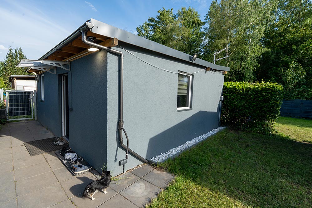 Schwarzweisse Katze blickt neugierig neben grauem Haus mit Holzvordach