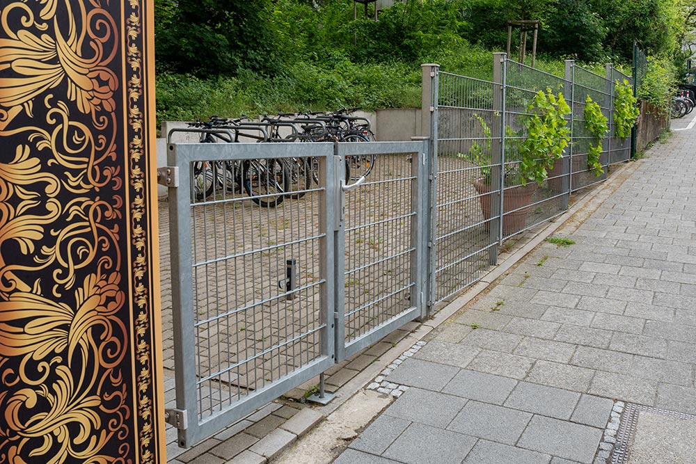 Graues Metalltor neben gold schwarzem Musterzaun vor Fahrrädern und Grün