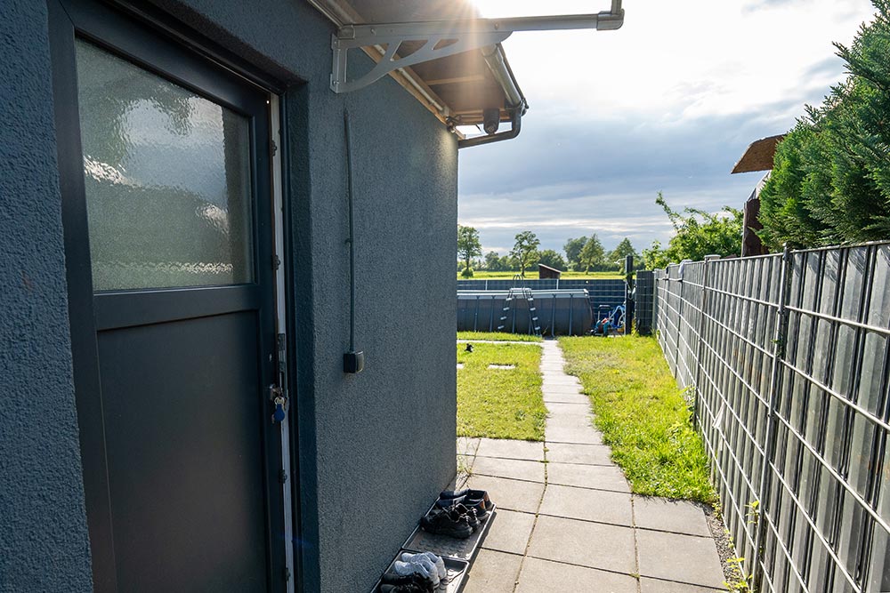 Hausgang mit grauer Tür links und sonnigem Weg zum Garten und Zaun rechts
