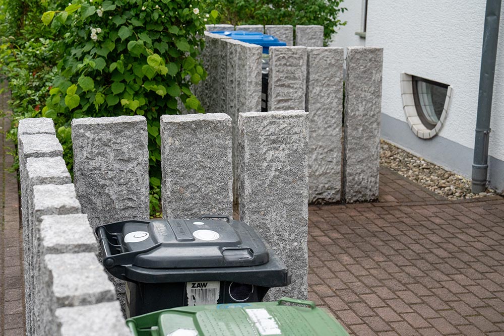 Reihe grauer Steinpfeiler neben Mülltonnen am Pflasterweg, ruhig und kantig