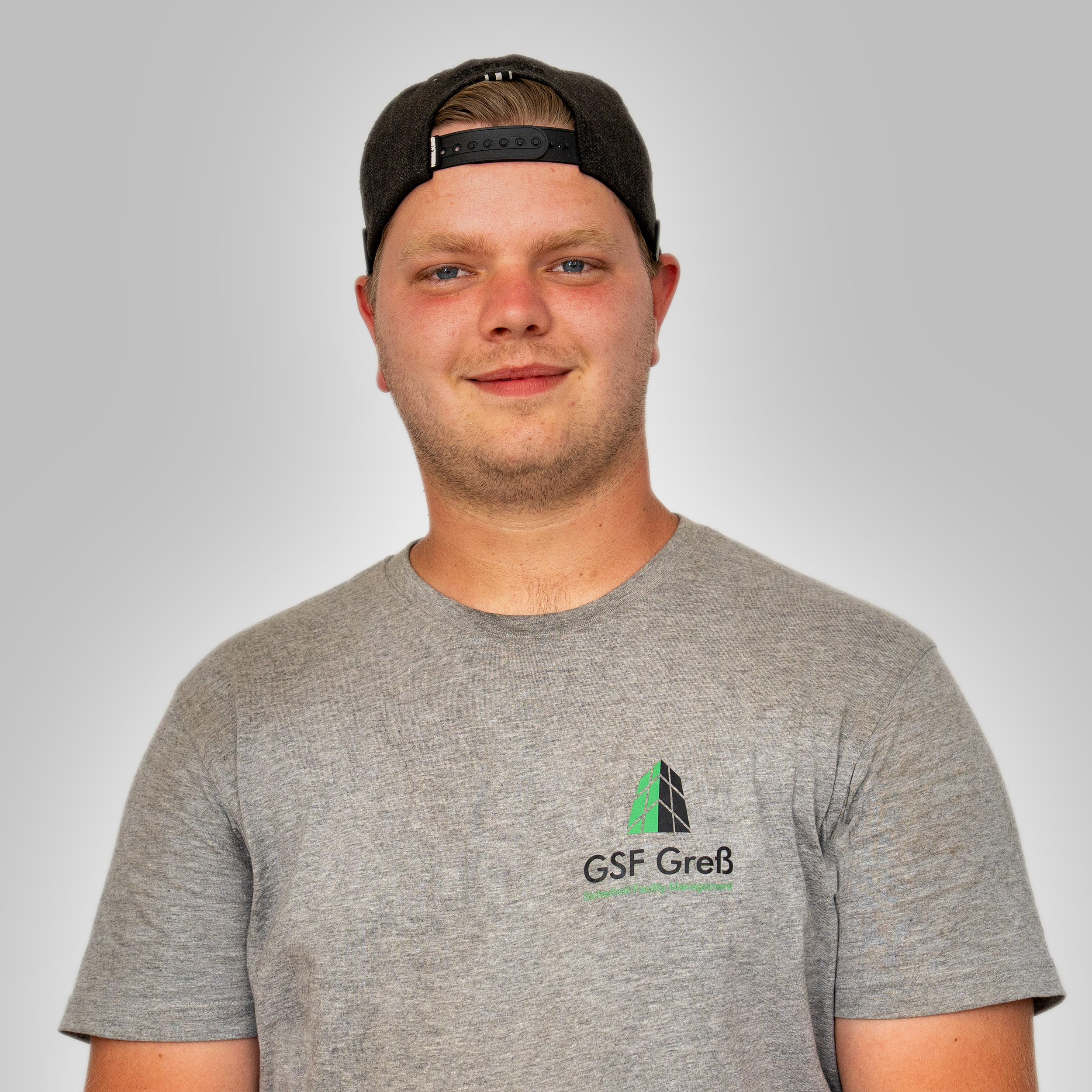 Junger Mann laechelt, schwarze Kappe rueckwaerts, graues Shirt mit Logo