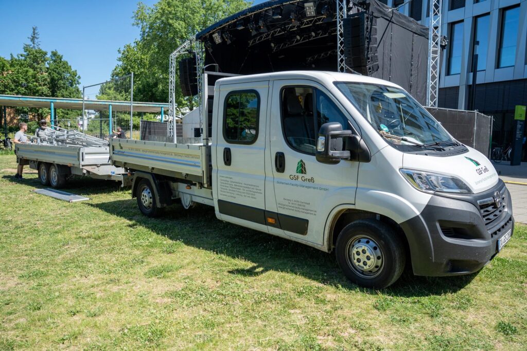 Weisser Pritschenwagen mit Anhaenger vor Buehne auf Wiese beim Aufbau
